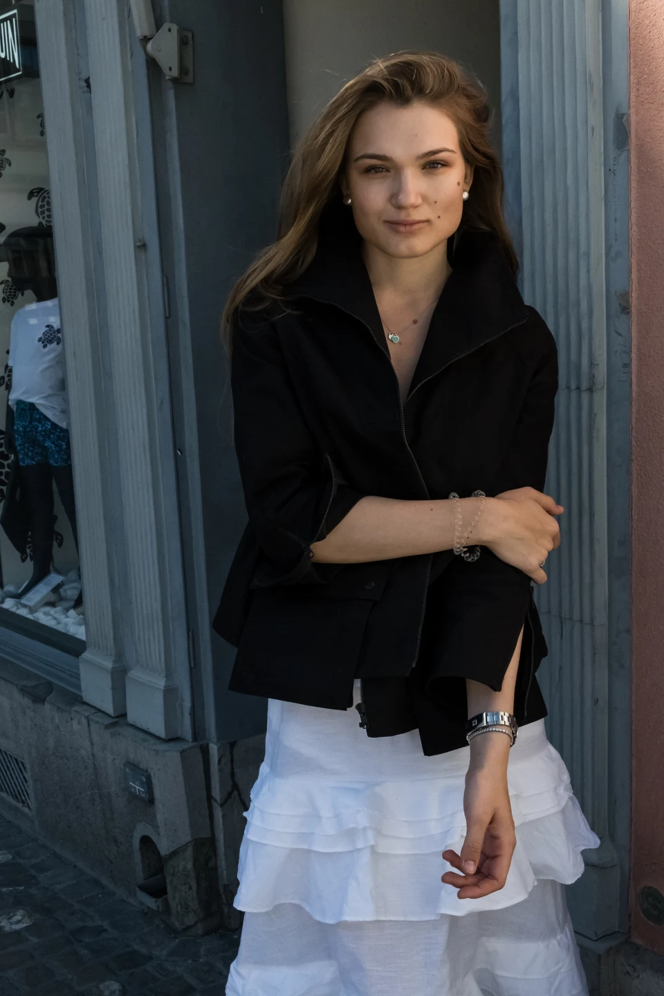 Fashion portrait in a black jacket and white skirt, framed by a blue doorway.