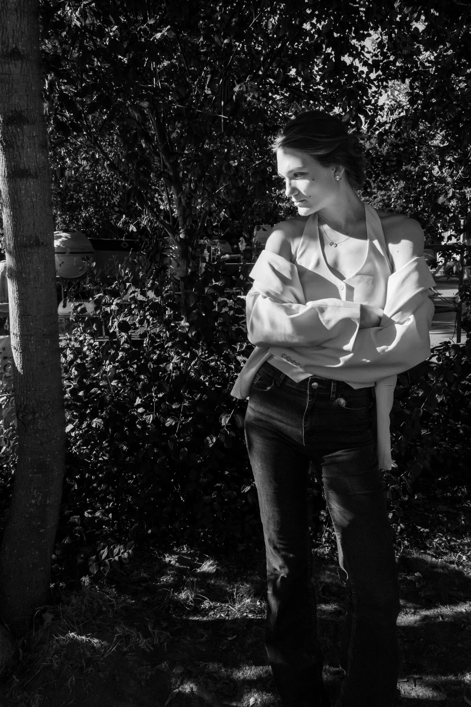 Monochrome fashion portrait in a pale blouse beside dense foliage.