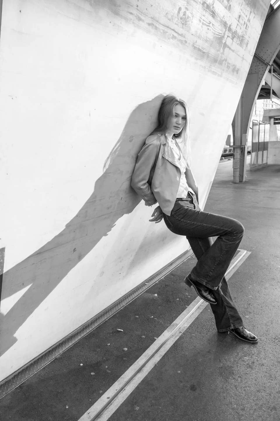 Full-length monochrome fashion portrait against a curved wall with a diagonal shadow.
