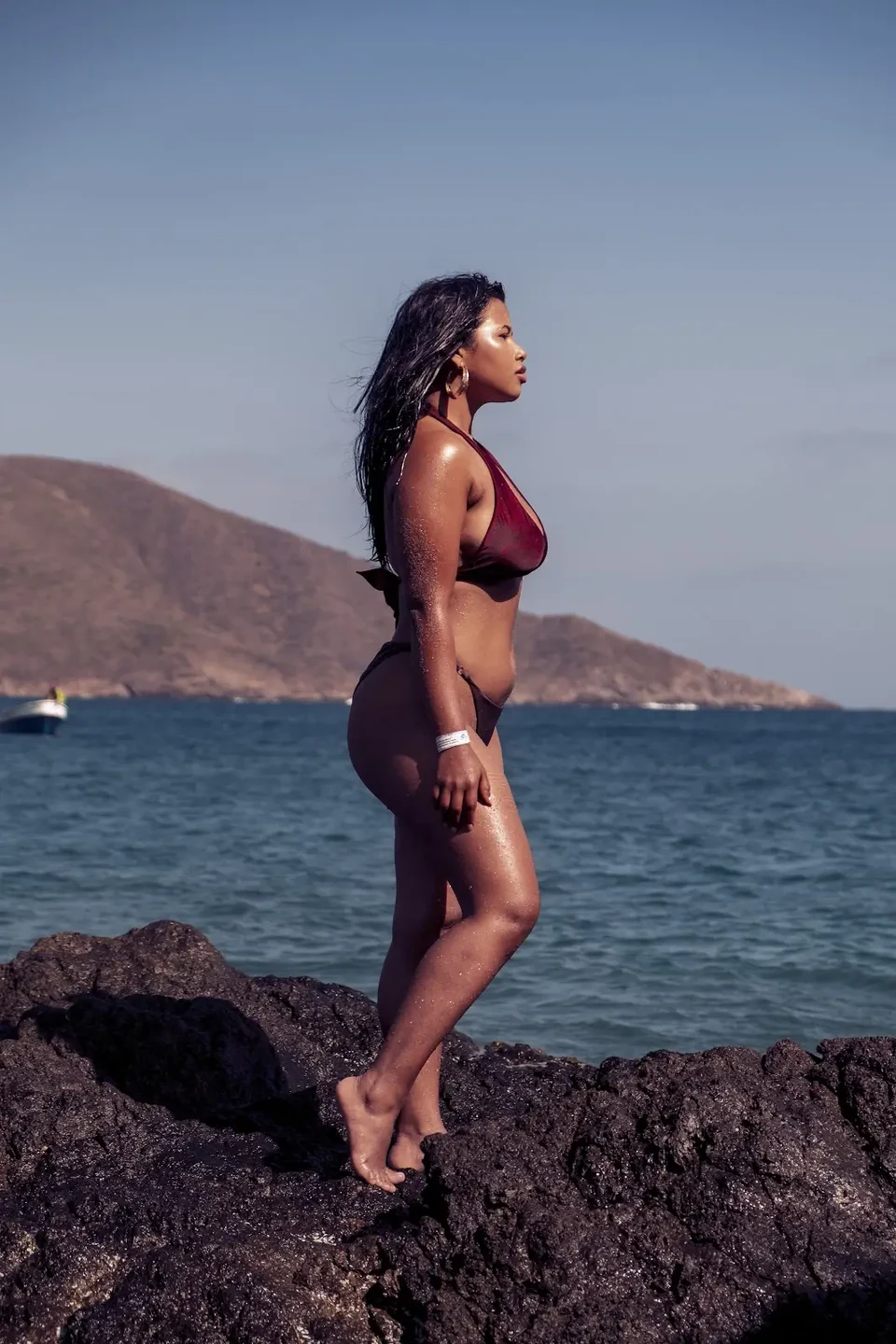 A model in burgundy swimwear standing on coastal rocks, with blue water and mountains behind her.